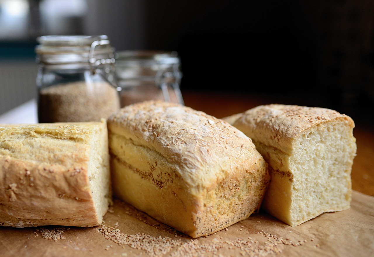 Three fresh loaves of lightly golden, yeast bread dusted with sesame seeds, resting on parchment paper with grain jars blurred in the background.